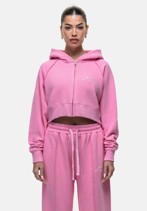 Felpa corta rosa con zip frontale, cappuccio e logo ricamato. Abbinata a pantaloni da tuta rosa coordinati con vita elasticizzata.