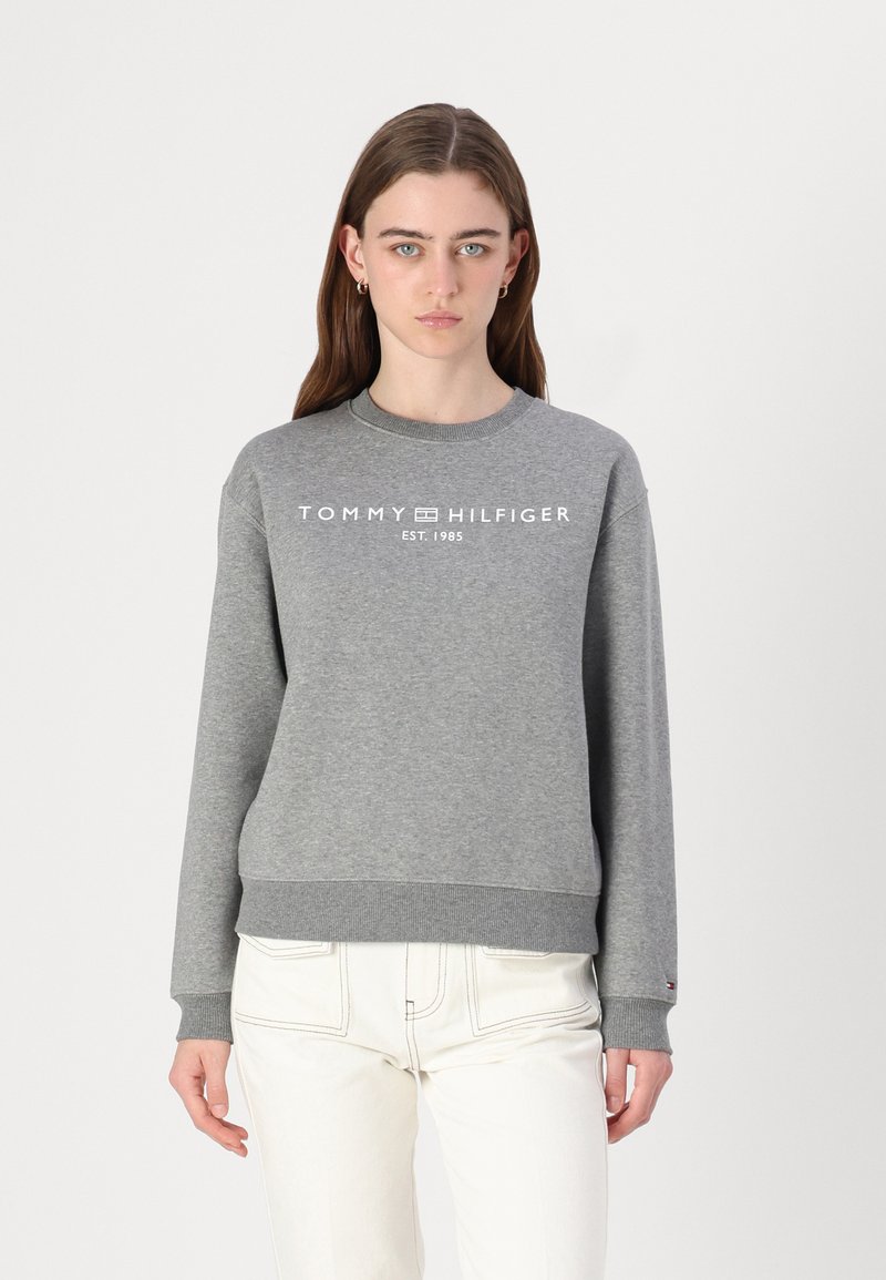 Pilkas sportinis megztinis su raukšlėtais rankogaliais ir apatine dalimi, priekyje su baltu "TOMMY HILFIGER" logotipu ir tekstu "EST. 1985". Lygus paviršius.