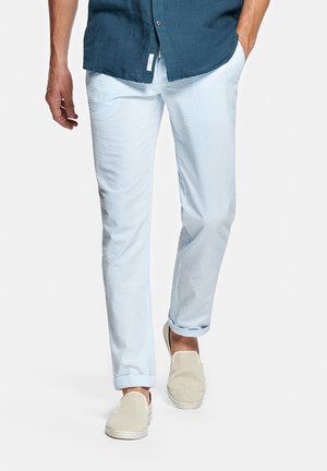 Homme marchant, portant un pantalon à rayures bleu clair et blanc, une chemise bleu marine et des chaussures beiges perforées sans lacets, main dans la poche.