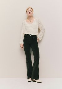 Blusa bianca con texture e maniche lunghe, abbinata a pantaloni neri a zampa d'elefante e ballerine beige. Presenta un collo quadrato e un dettaglio con fiocco in vita.