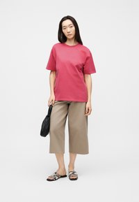 Γυναίκα που φοράει ροζ κοντομάνικο πουκάμισο, μπεζ παντελόνι φαρδύ μέχρι τον αστράγαλο, σανδάλια με σχέδιο φιδιού, κρατώντας μια μικρή μαύρη τσάντα.