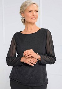 Schwarze Bluse mit langen durchsichtigen Ärmeln, Rundhals-Ausschnitt, gepunkteten Verzierungen und einer glatten Textur. Das Modell zeigt eine neutrale Pose mit verschränkten Händen.