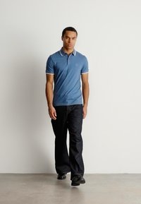 Polo shirt bleu avec des détails de col blanc, associé à un pantalon foncé et des chaussures noires. Le modèle se tient devant un fond neutre.
