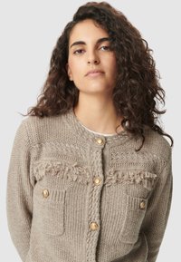 Donna con lunghi capelli ricci che indossa un cardigan lavorato a maglia beige con bottoni dorati e dettagli con frange su uno sfondo semplice.