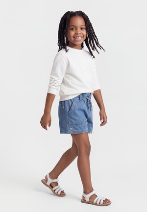 Usmievavé dievča s vrkočmi kráča, má na sebe bielu blúzku s dlhým rukávom, vyšívané rifľové šortky a biele sandále na jednofarebnom pozadí.