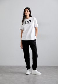 Hvit t-skjorte med korte ermer som har svart "EA7 Emporio Armani"-logo. Kombinert med svarte bukser og hvite sneakers, står på et grått gulv.