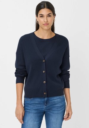 Marineblaue gerippte Strickjacke mit V-Ausschnitt, Knopfschluss und langen Ärmeln. Getragen über einem passenden Oberteil, kombiniert mit blauen Jeans.