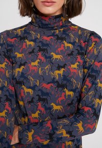 Mörkblå långärmad polotröja i tyg, med ett mönster av gula, röda och svarta hästsiluetter mot en strukturerad bakgrund.
