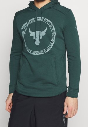 Homme portant un sweat à capuche vert foncé avec un motif circulaire de serpent entourant un symbole de tête de taureau sur la poitrine, associé à un pantalon noir.