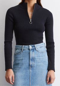 Frau in schwarzem, geripptem Langarmoberteil mit Reißverschluss und einem hellblauen, hochgeschnittenen Jeansrock, die vor einem schlichten Hintergrund steht.