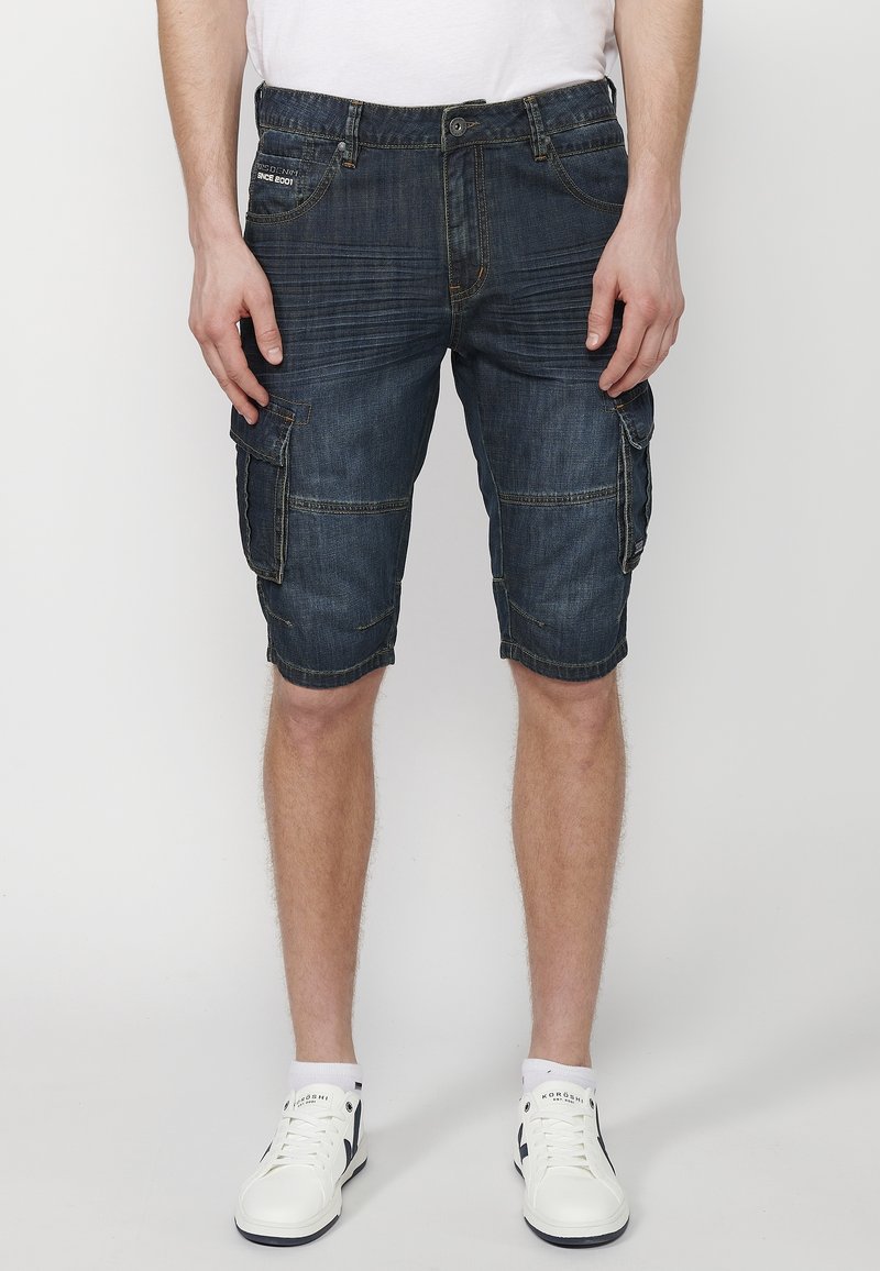 Shorts cargo in denim blu scuro con molteplici tasche, dettagli cuciti e una vestibilità ampia, abbinati a sneaker bianche e calzini in evidenza.