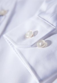Close-up van witte stof met een puntige kraag, twee ronde, doorzichtige knopen en zichtbare stiksels, wat de soepele textuur benadrukt.