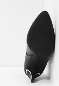 Svart högklackad sko med spetsig tå, slät läderöverdel och en karakteristisk böjd klack med silverdetaljer.