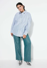Kobieta z ciemnymi włosami związanymi z tyłu, ubrana w oversize'ową białą i niebieską w paski koszulę, turkusowe błyszczące szerokie spodnie, srebrne botki na obcasie i kolczyki w kształcie kół.