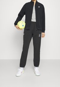 Chaqueta negra con cremallera y cuello alto, camiseta gris debajo, pantalones jogger negros, sosteniendo un balón de fútbol amarillo y zapatillas blancas con detalles de color.