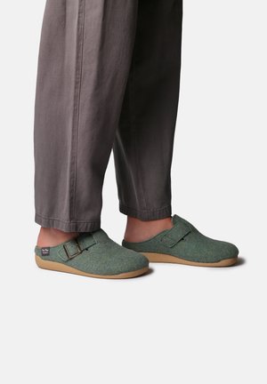 Sabots en feutre vert avec une sangle avant et une boucle en métal. Semelle légère beige et dessus lisse et texturé. Associés à un pantalon gris ample.