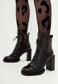 Botines negros de cuero, con tacón grueso, cierre de cordones en la parte frontal, cremallera lateral y detalles metálicos en la correa y la punta.