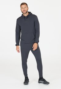 Dunkelgraues Sportset bestehend aus einem langärmeligen Oberteil mit Kapuze und Vordertasche sowie passenden schmal zulaufenden Jogginghosen. Strukturiertes Material, körpernahe Passform.