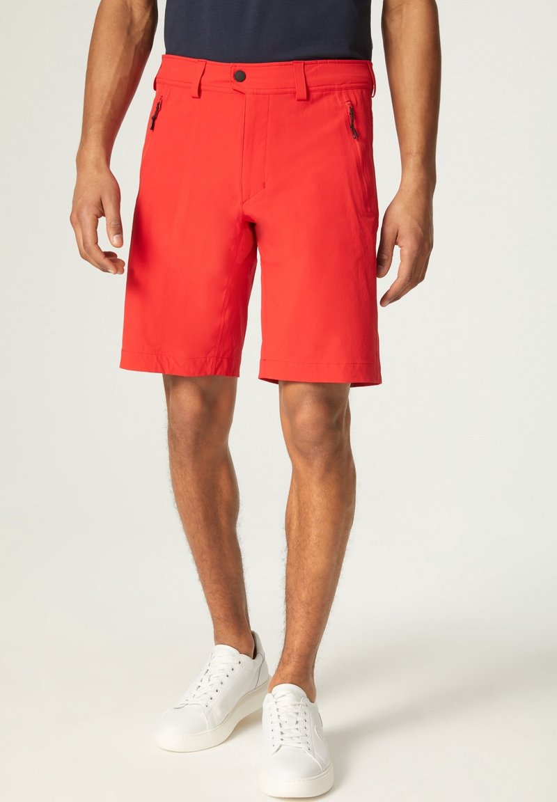 Shorts pour homme rouge vif arrivant aux genoux, avec poches zippées à l'avant, portés avec des baskets blanches et une chemise sombre.