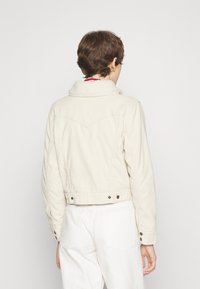 Beige kort jacka med fårskinnskrage, strukturerade axlar och sömnadsdetaljer. Ryggdelen har en dekorativ yoke och tryckknappsstängningar.