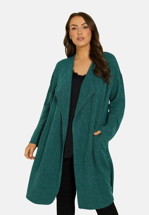 Frau mit langen braunen Haaren, die eine türkisfarbene offene Strickjacke über einem schwarzen Oberteil und schwarzen Hosen trägt, steht vor einem einfarbigen Hintergrund.