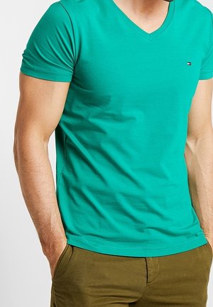 Mężczyzna ubrany w dopasowaną, oliwkowozieloną koszulkę z krótkim rękawem i dekoltem w serek oraz oliwkowe zielone spodnie, ze złożonymi rękami w kieszeniach, widziany od szyi do bioder.