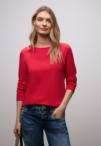 Vrouw met lang blond haar, gekleed in een rood shirt met lange mouwen en een blauwe spijkerbroek, staat met één hand in haar zak en de draagriem van een tas in haar andere hand.