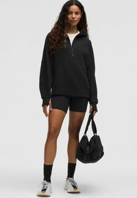 Czarny pulower z kapturem z półsuwałem i przednią kieszenią, w połączeniu z czarnymi szortami rowerowymi, czarnymi skarpetkami do kostek i białymi trampkami, trzymający czarną torbę.