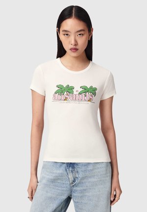 Jeune femme portant un t-shirt blanc avec des palmiers verts et le texte rose "all saints", assorti à un jean bleu clair, debout devant un fond gris.