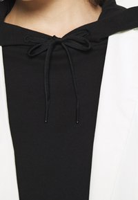 Černá mikina s kulatým výstřihem, stahováním na šňůrku a texturovanou látkou, doplněná bílým svrchním oděvem.