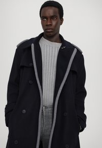 Cappotto di lana blu navy con dettagli grigi, bottoni doppi e colletto strutturato. Indossato sopra un maglione grigio a coste e jeans chiari.