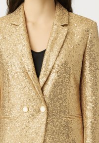 Blazer dorato in paillettes con tessuto testurizzato e design sartoriale, rever a rotonda e due bottoni bianchi sul davanti.