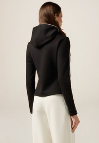 Giacca nera con cappuccio dal design aderente, con piping bianco lungo le cuciture, texture liscia e lunghezza cropped, abbinata a pantaloni bianchi.