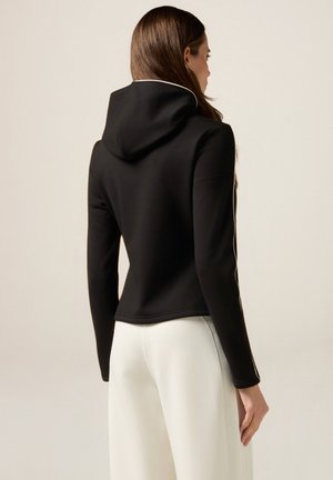 Giacca nera con cappuccio dal design aderente, con piping bianco lungo le cuciture, texture liscia e lunghezza cropped, abbinata a pantaloni bianchi.