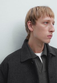 Dark gray wool coat with a lapel collar over a gray knit sweater, layered above a white t-shirt, showcasing a minimalistic design.