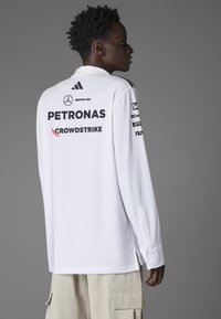 White long-sleeve shirt featuring black logos and text, including "PETRONAS" and "CROWDSTRIKE," with buttoned cuffs and side slits.