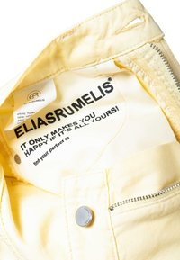 Tissu couleur vanille avec une fermeture éclair, un bouton-pression et une étiquette portant l'inscription "ELIASRUMELIS" ainsi que la phrase "Ça ne te rend heureux que si c'est entièrement à toi !"