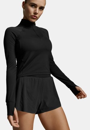 Kobieta ubrana w czarną sportową bluzę z długim rękawem i otworami na kciuki oraz dopasowane czarne spodenki, stojąca na jasnym, jednolitym tle.