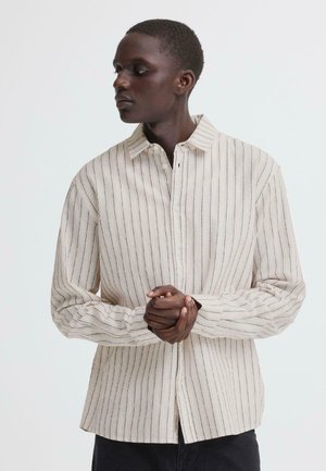 Personne portant une chemise beige à manches longues avec de fines rayures verticales noires, debout les mains jointes, regardant sur le côté.