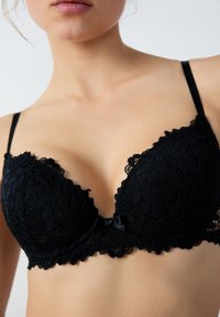Schwarzer Spitzen-BH mit tiefem Ausschnitt. Blumenmuster, gewellter Saum und ein zentraler Seidenbogenakzent. Verstellbare Träger und vorgeformte Cups.