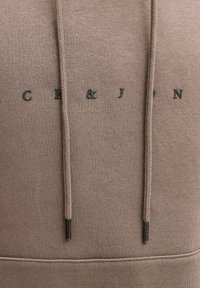 Pruun kapuutsiga dressipluus, millel on mustad metallist paelakolmnurgad ja esiküljel tumedates toonides trükitud logo "CK & J". Pehme kangatekstuuri.