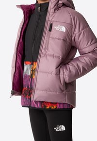 Osoba ubrana w jasnofioletową kurtkę puchową The North Face na kolorowym polarze oraz czarne legginsy The North Face.