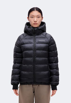 Un mannequin est de face, vêtu d’une doudoune noire matelassée à capuche Peak Performance et d’un pantalon taupe, sur un fond uni.
