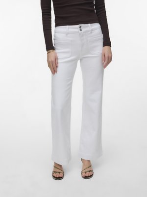 Kvinde iført hvide bukser med høj talje og vide ben med forlommer kombineret med sort langærmet top og sorte sandaler.