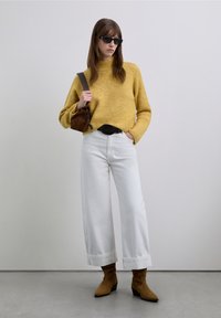 Pull en tricot jaune, jean blanc à jambes larges, bottines en daim marron et sac marron. Accessoires : lunettes de soleil noires. Fond minimaliste.