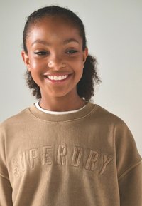 Brun sweatshirt med ribbad halsringning, med "SUPERDRY" präglat i matchande färg. Mjuk textur och avslappnad passform. Vit t-shirt synlig.