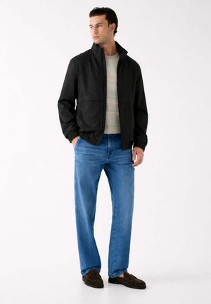 Uomo con giacca nera con zip, maglione beige, jeans blu e mocassini marrone scuro, in piedi con le mani nelle tasche e che guarda verso sinistra.