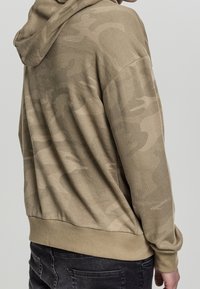 Bežová mikina s texturovaným maskáčovým vzorem, s kapucí a vroubkovanými manžetami, v kombinaci s tmavými džíny pro kontrast.