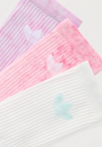 Tre par sokker: lilla, rosa og hvite. Hver har vertikal ribbestrikk og en diskret løvlogo i kontrasterende farger på ankelen.