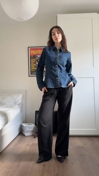 Jauna sieviete stāv guļamistabā, valkājot džinsa pogājamu kreklu, melnas plaša piegriezuma bikses un melnas smailas kurpes, aiz viņas ir Pulp Fiction plakāts.